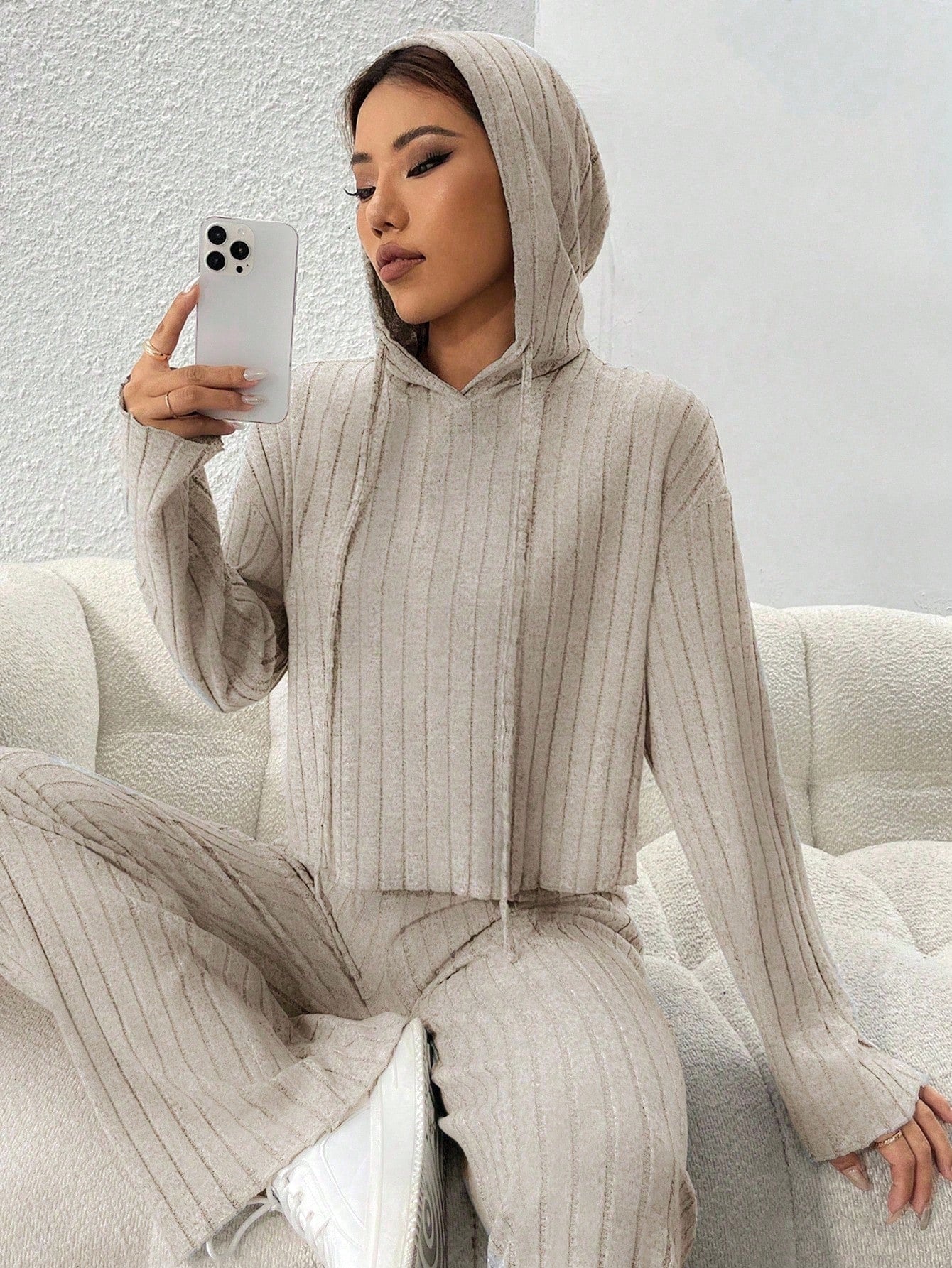 Woman in a beige ribbed hoodie and pants sitting on a white couch, holding a phone. Nushes