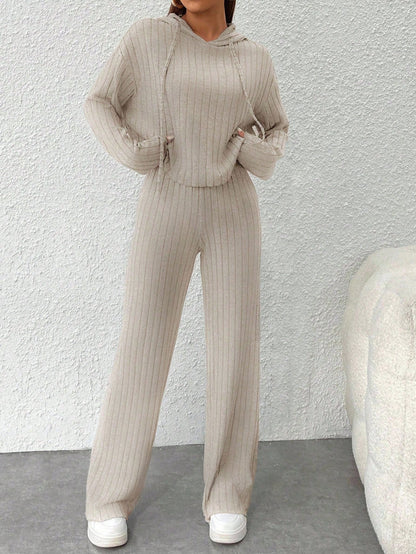 Person wearing a beige ribbed knit outfit with a matching hoodie and pants against a white wall. Nushes