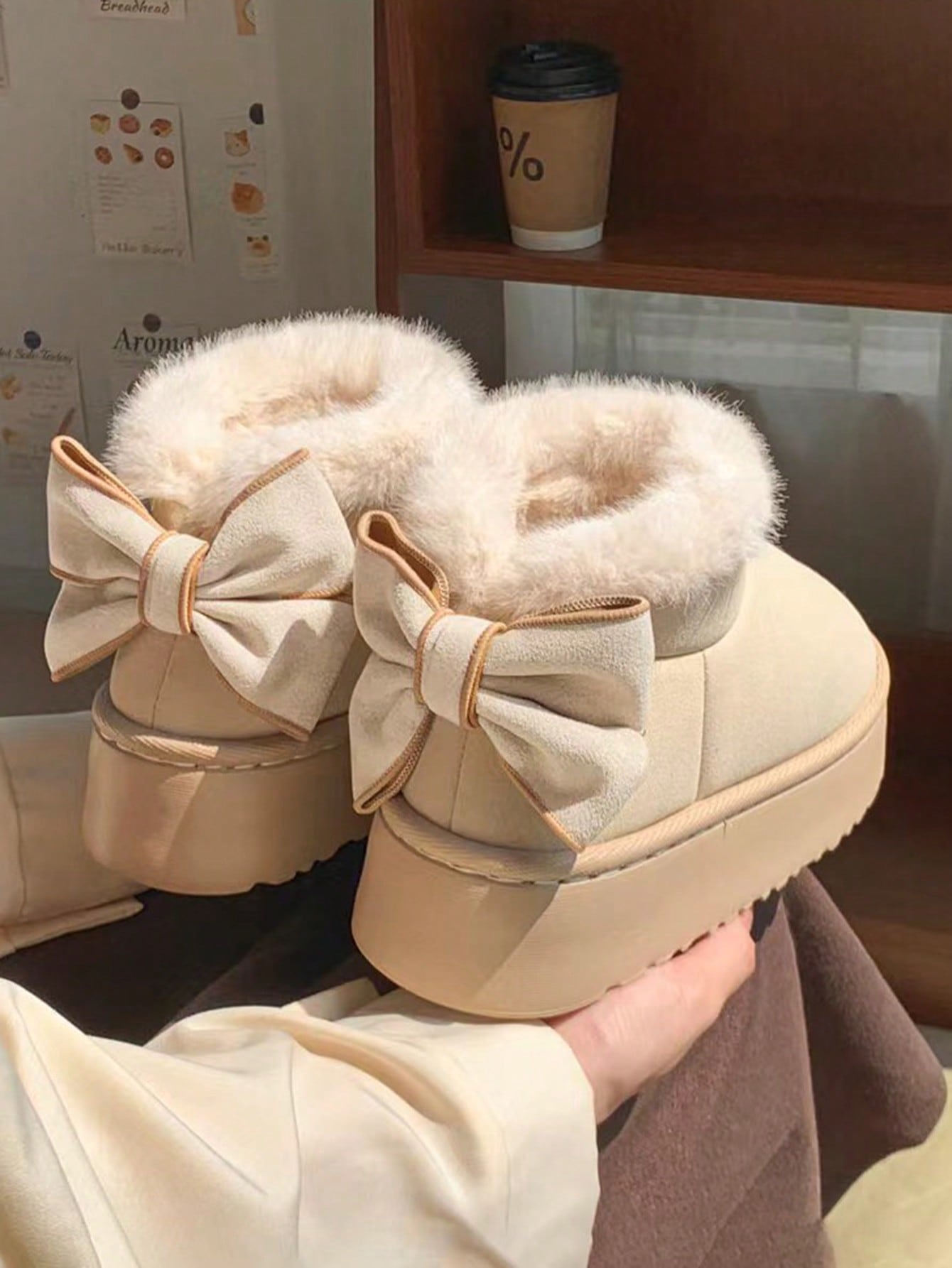 Beige snow boots with large bows held by a hand, with a blurred background.