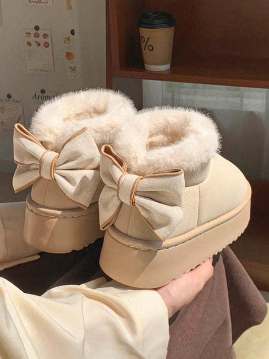 Beige snow boots with large bows held by a hand, with a blurred background.