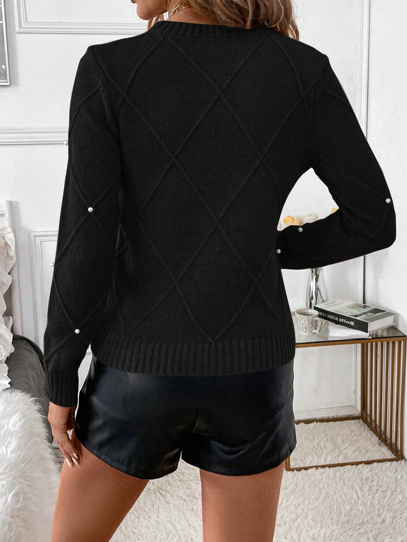 Person wearing a black sweater with pearl embellishments and black shorts in an indoor setting. Nushes