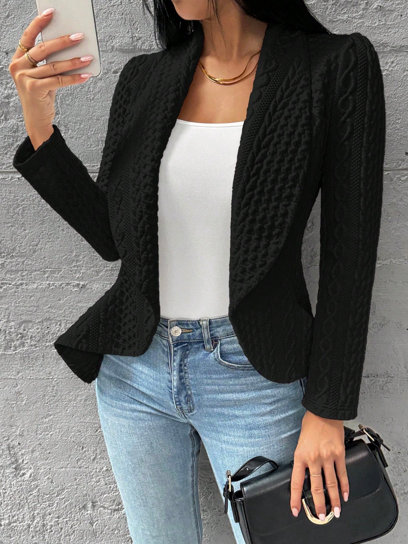 Person wearing a black textured blazer, white top, blue jeans, and holding a black handbag against a gray wall. Nushes 