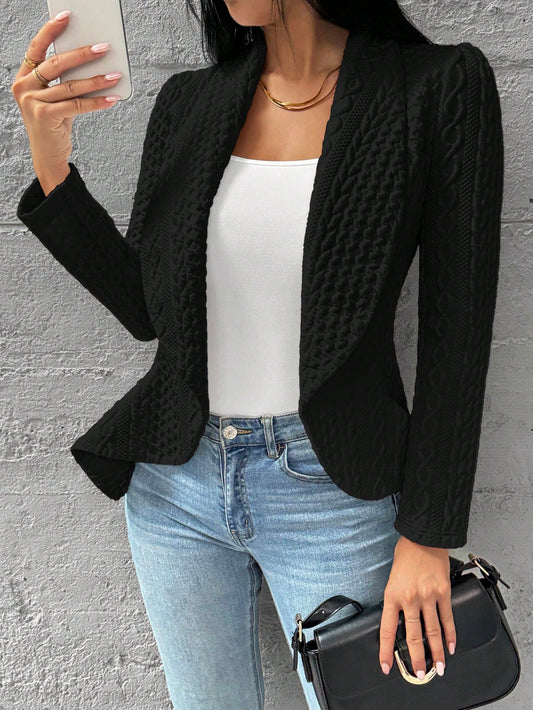 Person wearing a black textured blazer, white top, blue jeans, and holding a black handbag against a gray wall. Nushes 