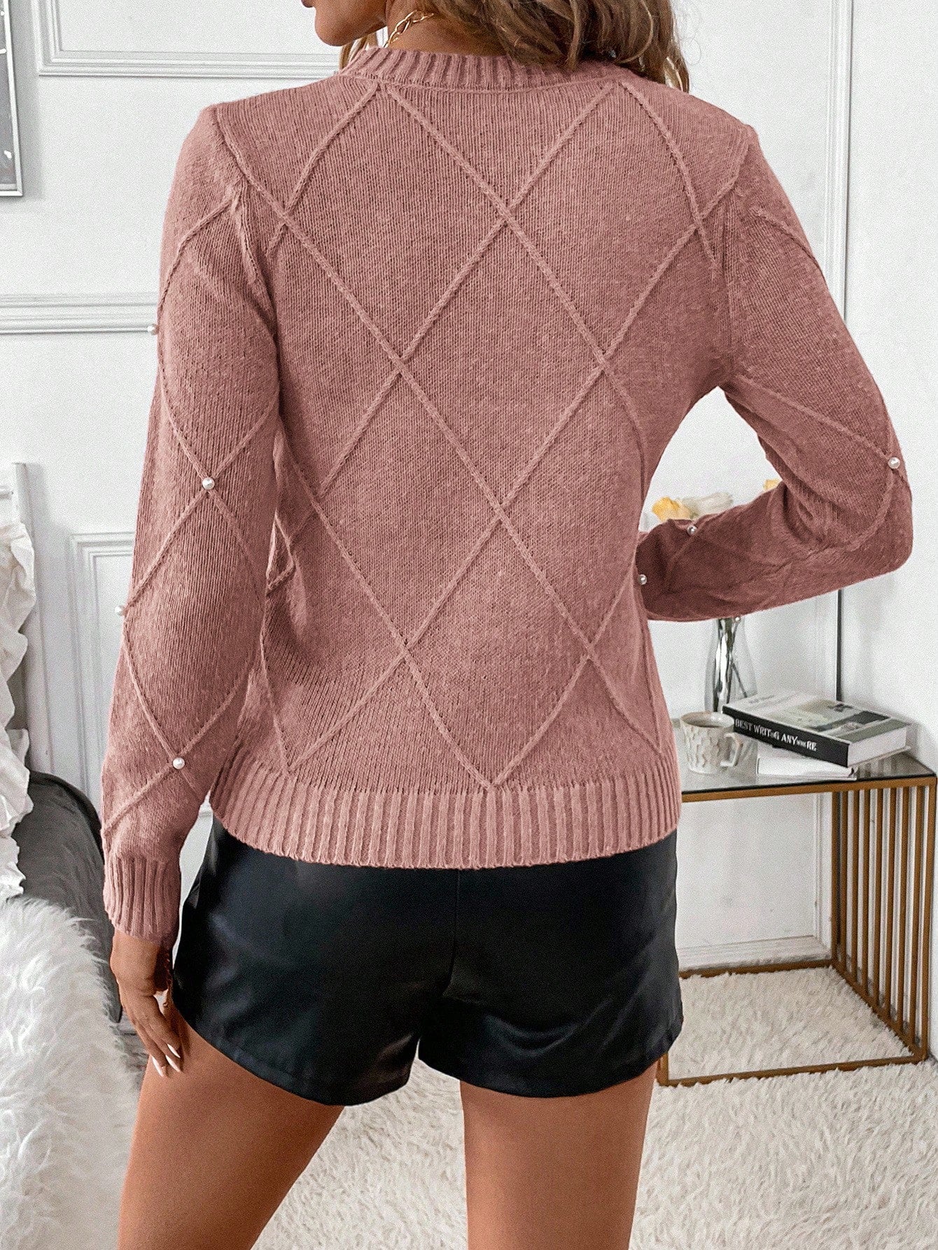 Person wearing a pink sweater with a diamond pattern and black leather shorts in a bedroom setting Nushes