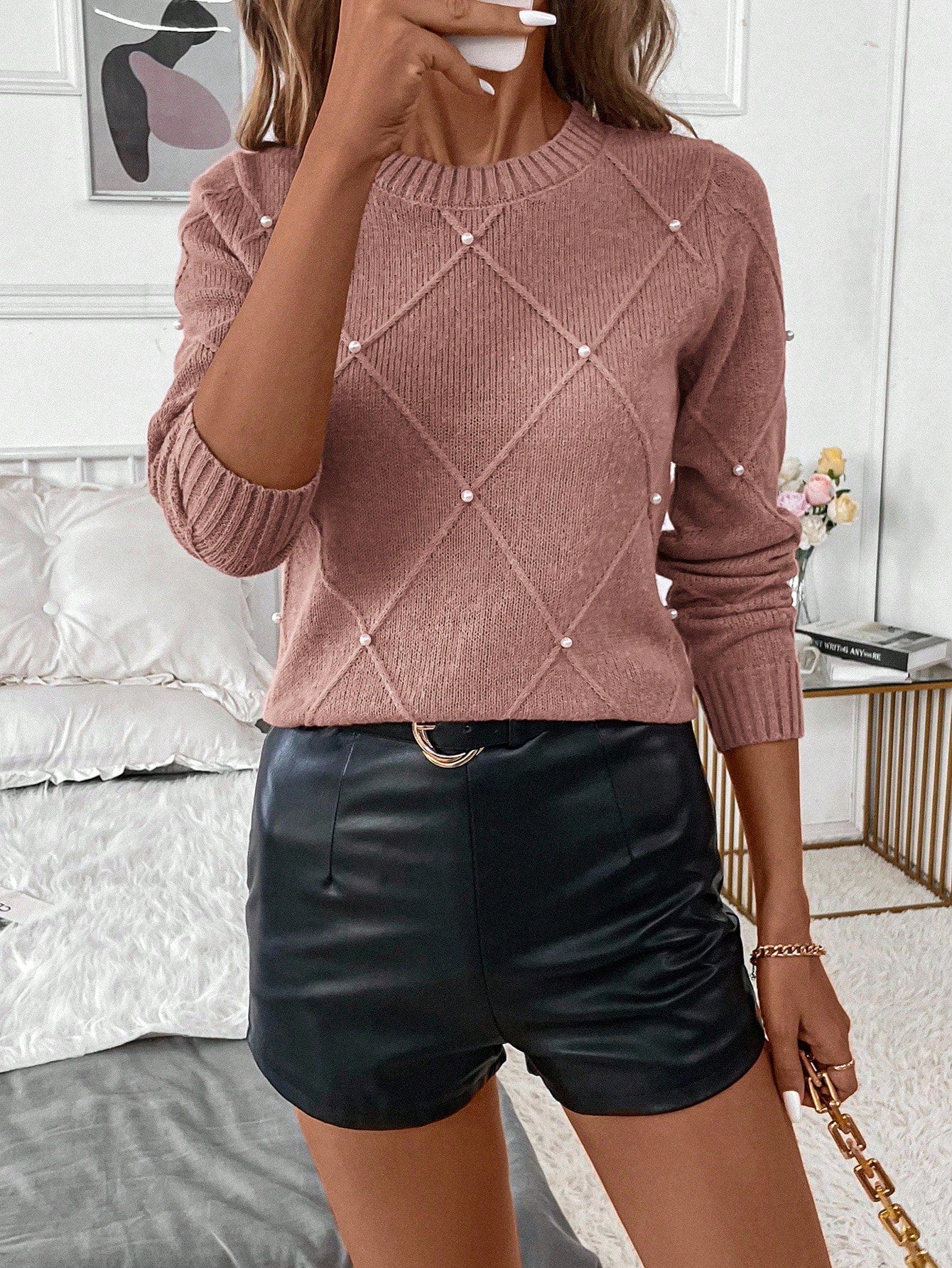 Person wearing a pink sweater with a diamond pattern and black leather shorts in a bedroom setting Nushes