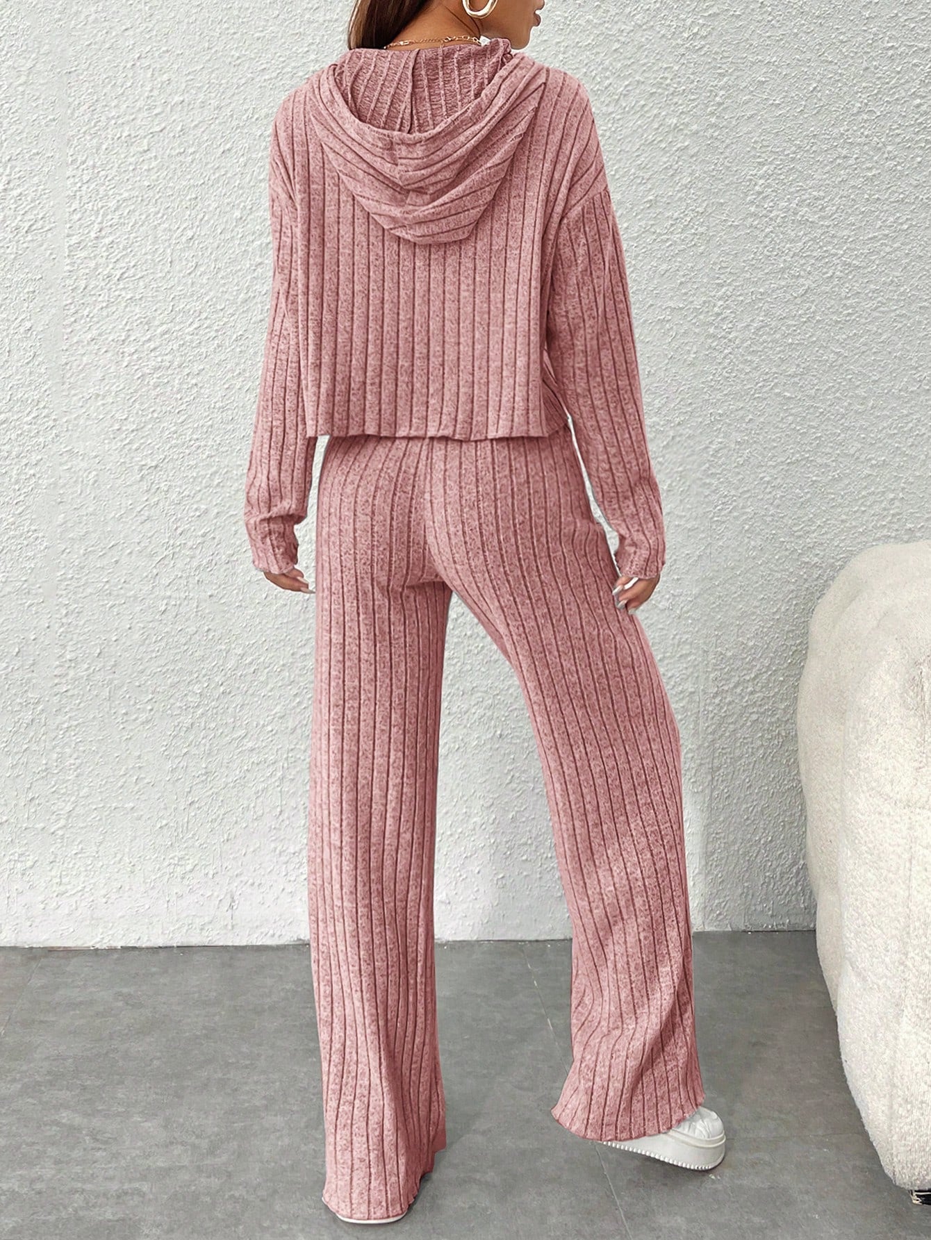 Person wearing a pink ribbed hoodie and pants set against a white wall.Nushes