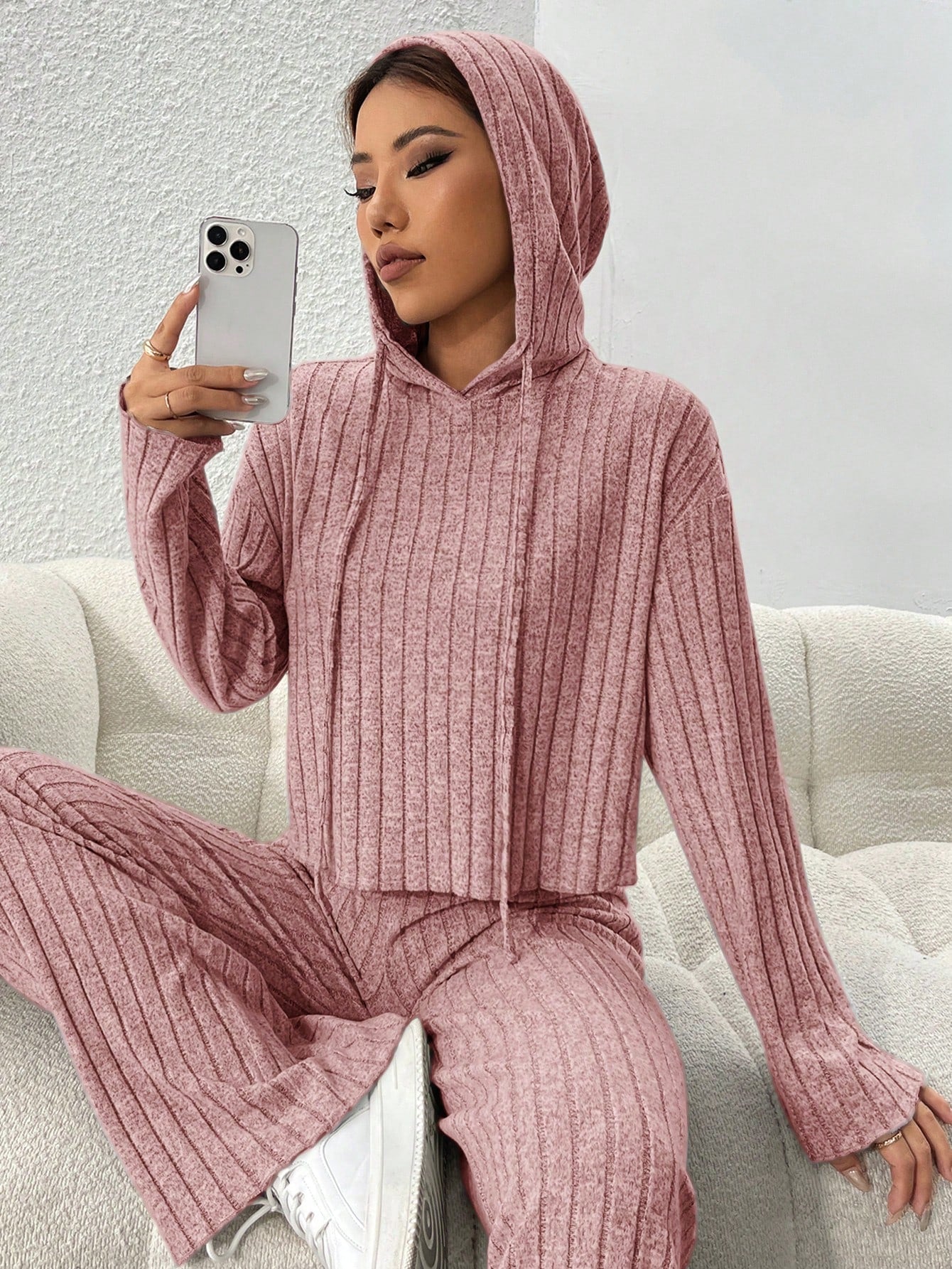 Person wearing a pink ribbed hoodie and pants sitting on a white couch. Nushes