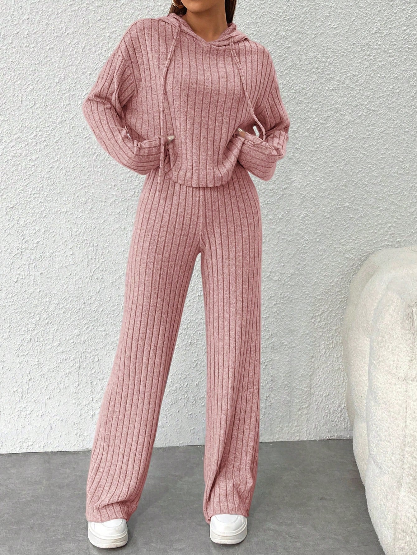 Person wearing a pink ribbed sweater and pants set against a light gray wall. Nushes