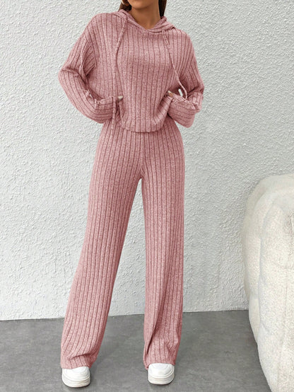 Person wearing a pink ribbed sweater and pants set against a light gray wall. Nushes