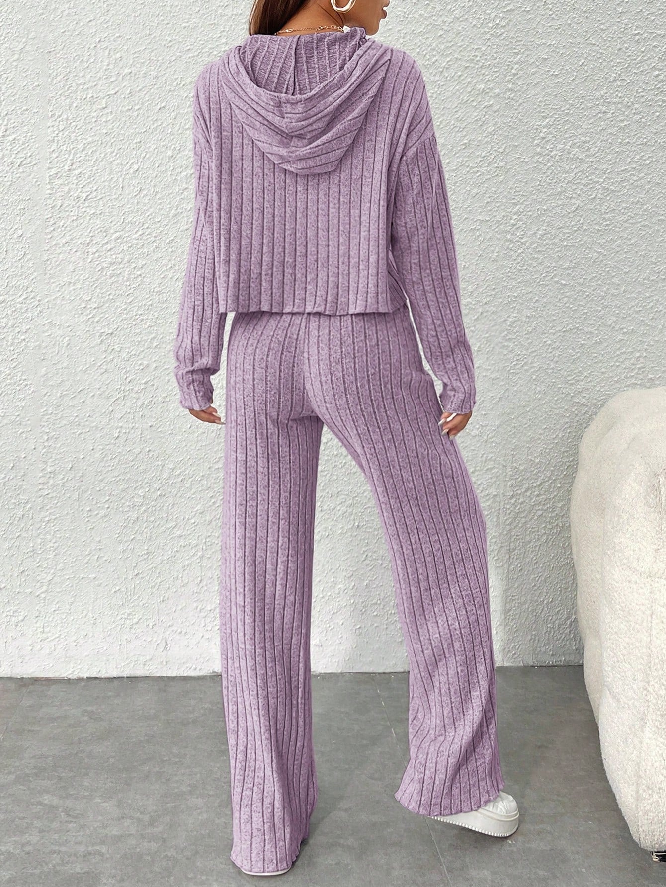Person wearing a purple ribbed hoodie and pants set against a white wall.Nushes