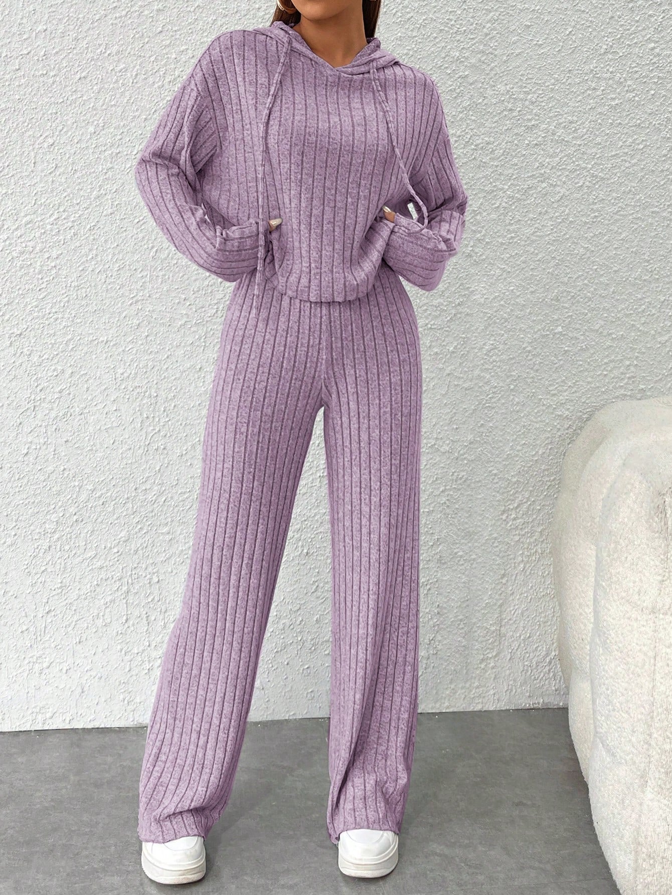 Person wearing a purple ribbed sweater and pants set against a white wall.Nushes