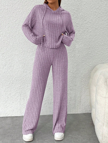 Person wearing a purple ribbed sweater and pants set against a white wall.Nushes